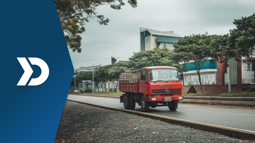 A red delivery truck driving on an urban road, surrounded by trees and modern buildings, symbolizing efficient logistics and fleet management.